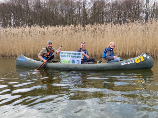 „We stand with the World‘s Rangers“ – Kanutour mit Rangern im Müritz-Nationalpark. Foto: Patric Heintz. Auf dem Bild zu sehen sind: 3 Ranger im Kanu vor Schilf mit einem Banner "We stand with the World's Rangers"