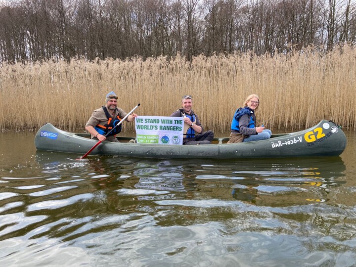 „We stand with the World‘s Rangers“ – Kanutour mit Rangern im Müritz-Nationalpark. Foto: Patric Heintz. Auf dem Bild zu sehen sind: 3 Ranger im Kanu vor Schilf mit einem Banner "We stand with the World's Rangers"