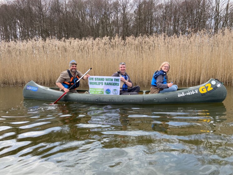 „We stand with the World‘s Rangers“ – Kanutour mit Rangern im Müritz-Nationalpark. Foto: Patric Heintz. Auf dem Bild zu sehen sind: 3 Ranger im Kanu vor Schilf mit einem Banner "We stand with the World's Rangers" „We stand with the World‘s Rangers“ – Kanutour mit Rangern im Müritz-Nationalpark. Foto: Patric Heintz. Auf dem Bild zu sehen sind: 3 Ranger im Kanu vor Schilf mit einem Banner "We stand with the World's Rangers"