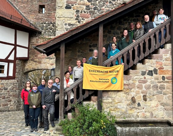 Rund 20 Erwachsene stehen auf einer Holztreppe, eingefasst in eine alte Mauer, vor sich ein Banner "Ehrensache Natur".